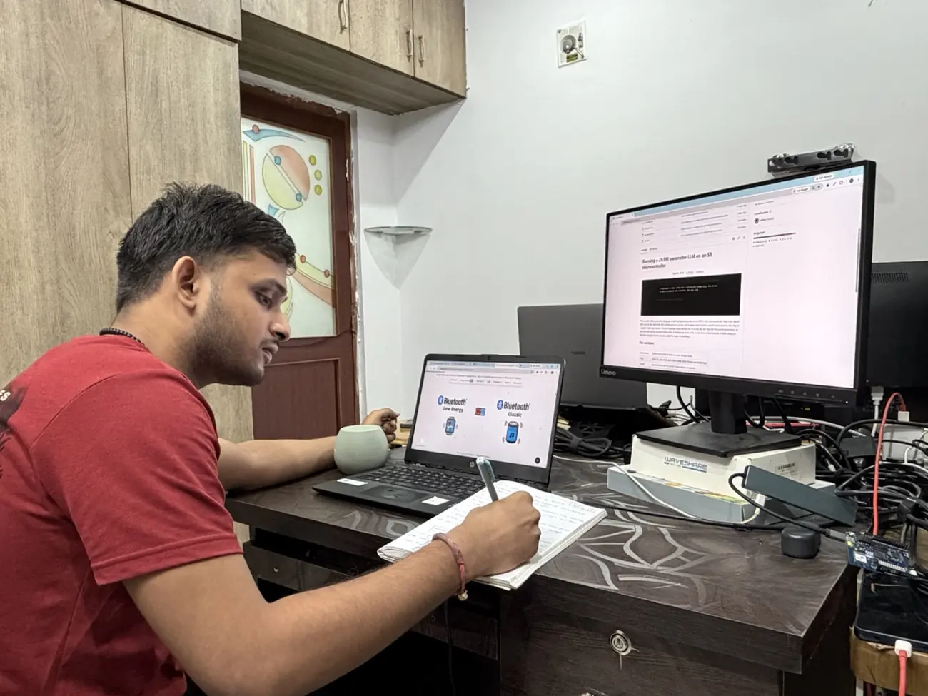 Harsha Vardhan Patri at a desk writing notes by hand, with a laptop showing Bluetooth Low Energy and Bluetooth Classic side by side and a development board on the bench.