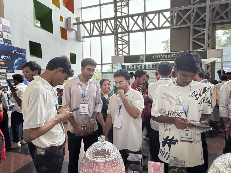 Visitors at the booth looking at phones and Dharanova cards while the scoreboard runs behind them.