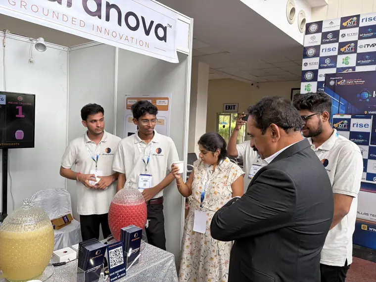A Dharanova engineer handing a small paper cup of juice to a visitor at the booth.