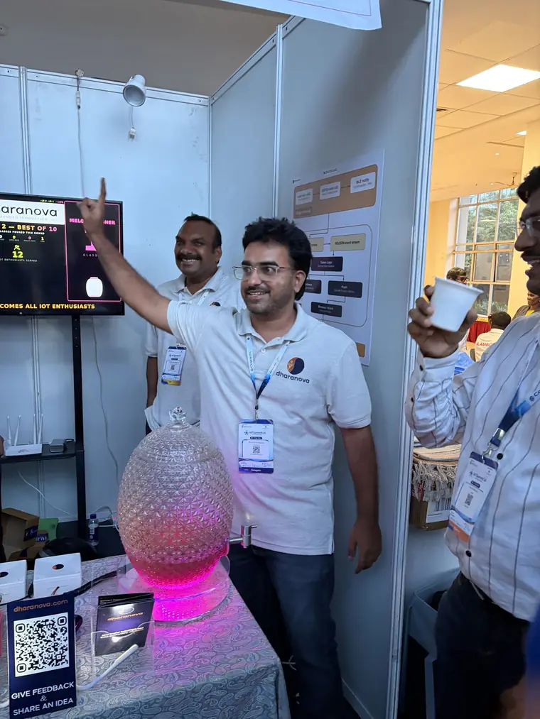 Nanupatruni Mahesh Kumar pointing up at the live scoreboard at the Dharanova booth, a lit juice jar on the table in front of him.