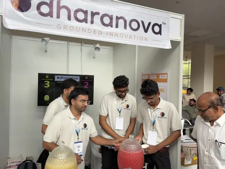 Nanupatruni Mahesh Kumar and Ramu Madada helping a visitor to a cup of juice at the Dharanova booth, the scoreboard reading 3 to 2 behind them.