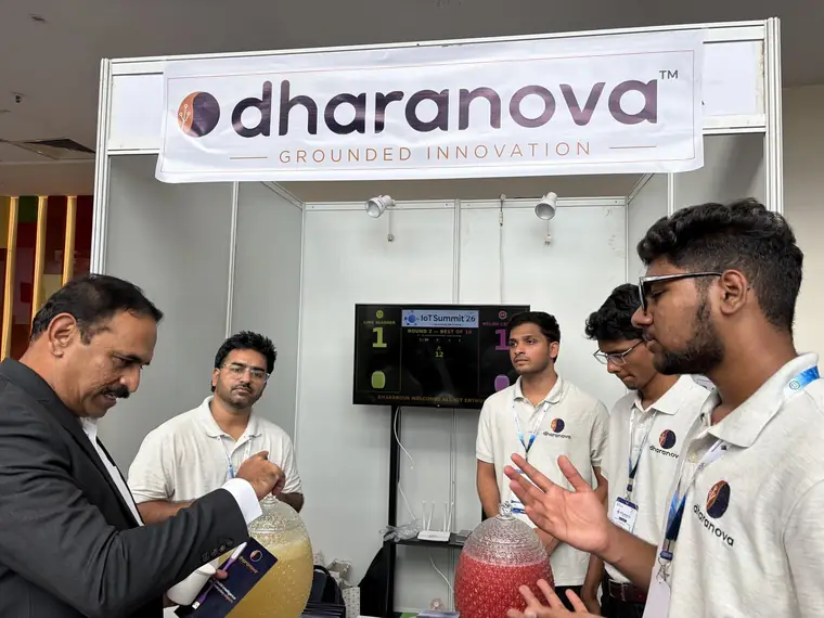A visitor in a suit talking to the Dharanova team beside the jars, with the scoreboard visible behind them.