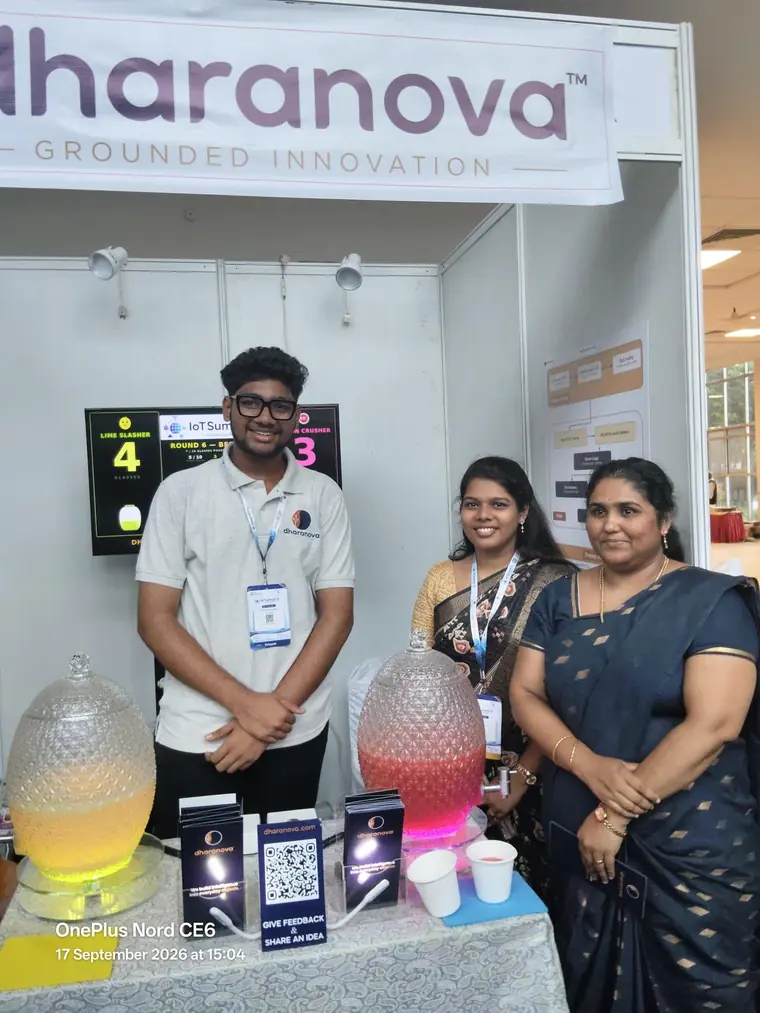 A Dharanova engineer standing with two visitors beside the jars, the feedback card on the table and the scoreboard behind showing round 6 with lime ahead 4 to 3.