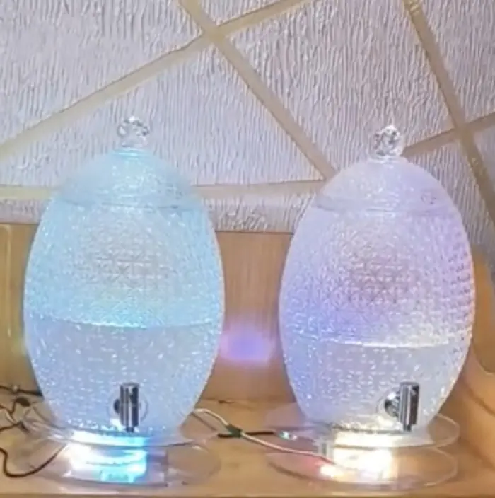 Two glass juice dispensers with taps, standing on lit platforms with cables running out of them.