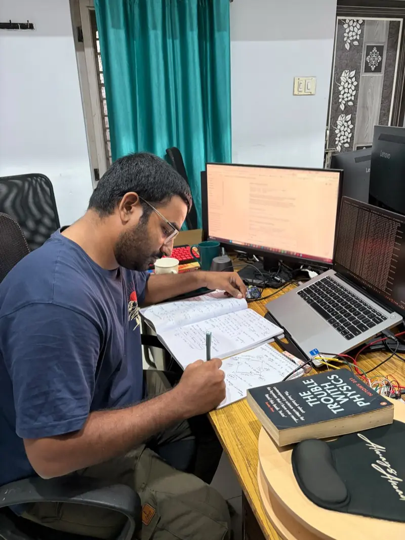 Nanupatruni Mahesh Kumar at a workbench, sketching a circuit diagram in a notebook, with a monitor of code, a laptop and jumper wires on the desk beside him.