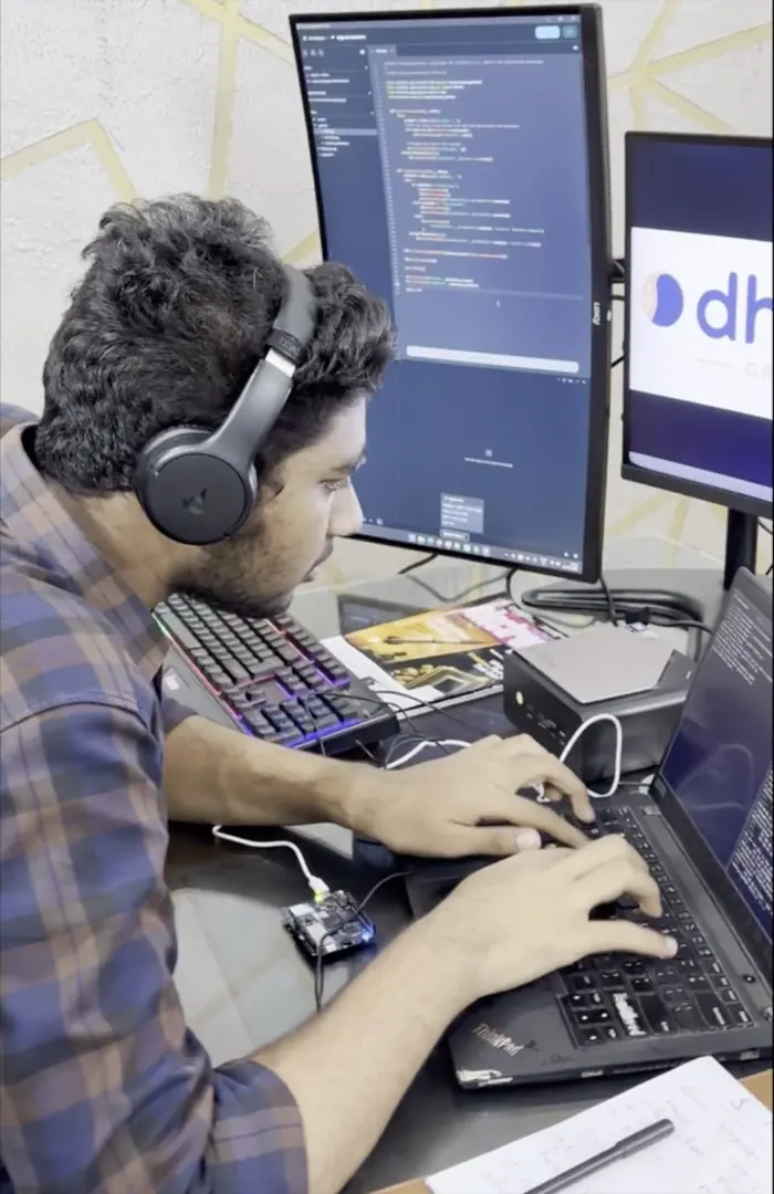 Ritesh Bonthalakoti wearing headphones, typing on a laptop with a small development board wired to it and a monitor of code behind.